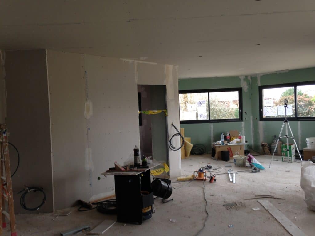 Pièce en pleine rénovation: murs et plafond en placo brut, fenêtres avec plans architecte. Outillage et matériaux de chantier au sol.