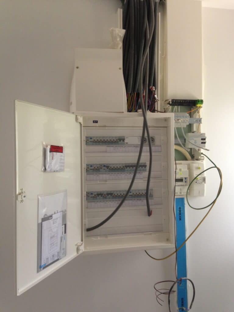 Tableau électrique mural ouvert montrant disjoncteurs, câbles gainés, DTI et composants réseau. Documents 'DOCUMENTS' et 'MICHAUD' visibles.