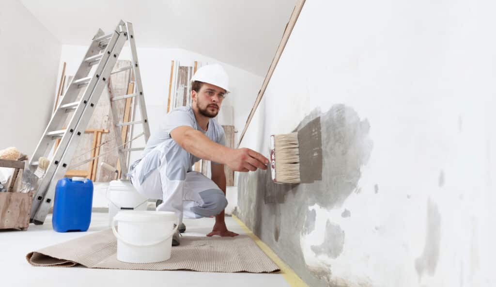 Homme en casque de chantier applique un primaire gris au pinceau sur un mur blanc. Chantier de rénovation avec échelles, outils et protections au sol.