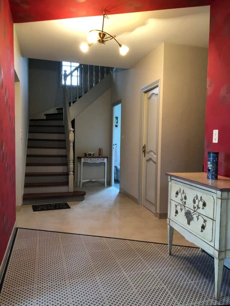 Entrée chaleureuse avec papier peint rouge, escalier en bois et commode vintage sous un luminaire moderne.